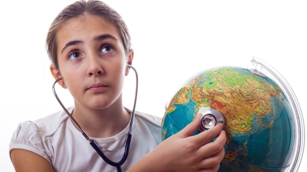 Menina segura globo mundial na mão e o escuta por meio de um estetoscópio. A imagem tem por objetivo ilustrar o tema da Saúde Planetária, que correlaciona a saúde ambiental com a humana. 